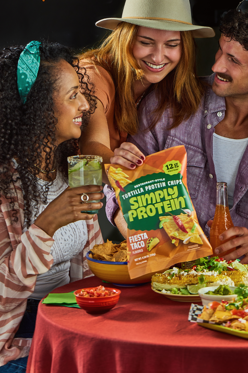 Three people at a table enjoying a meal with a bag of Fiesta Taco flavoured  SimplyProtein tortilla protein chips.