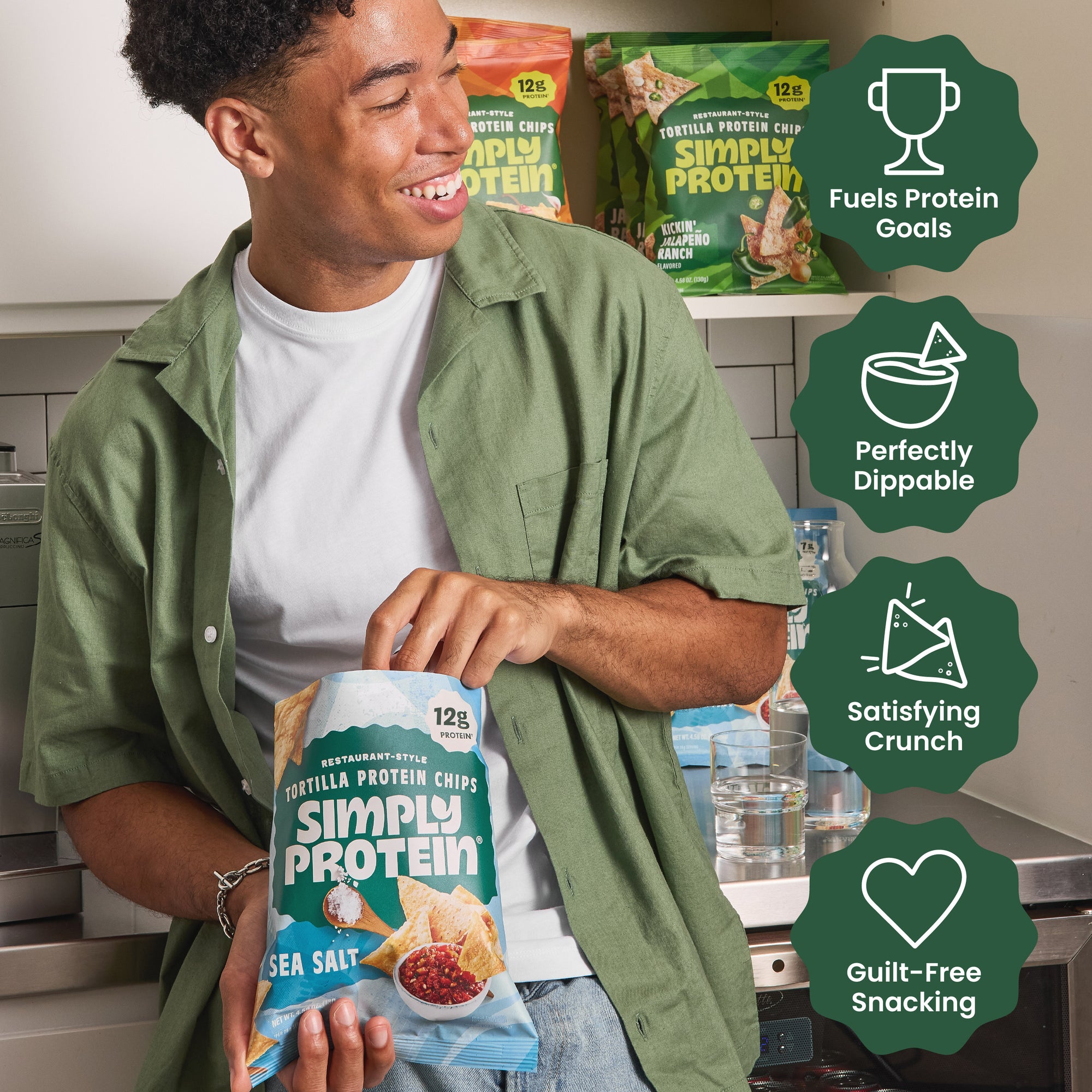 Person in a kitchen standing at a counter holding a package of Simply Protein protein tortilla chips with a cabinet behind them stocked with other flavored bags.